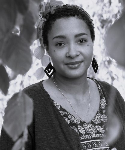 Lyrikerin Olaide Frank blickt freundlich in die Kamera. Sie ist eine Schwarze Frau mit kurzen Afrohaaren. Sie trägt eine Halskette, lange Ohrringe und ein gemustertes Oberteil